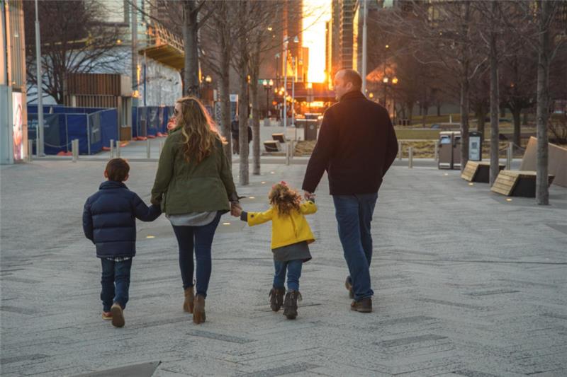 A family of four walks hand in hand down a city street at sunset. The warm glow casts long shadows, creating a serene, cheerful atmosphere.